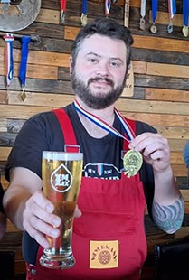 Man wearing a red apron holds up a glass of beer while displaying a medal around his neck.