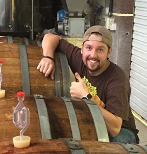 Man with backwards hat gives a thumbs up while leaning on beer barrels.