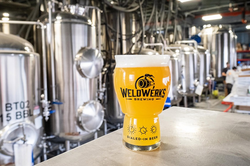 Beer in WeldWerks glass with a row of brewing equipment in the background.